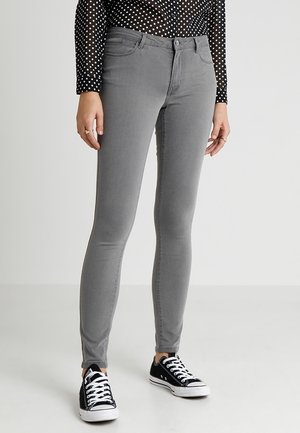 Kobieta ubrana w szare jeansy typu skinny, czarno-biały top w grochy z długim rękawem i czarne niskie trampki z białymi sznurówkami, stojąca.
