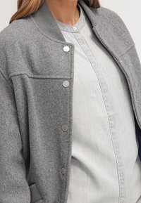 Bomber jacket grigio in misto lana con colletto a costine, bottoni a scatto e una finitura texture, indossato sopra una camicia chiara con bottoni.