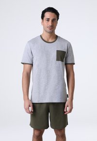 Maglietta grigia a maniche corte con dettagli verde scuro e taschino sul petto, abbinata a pantaloni corti verde scuro. Entrambi realizzati in tessuto morbido e leggero.