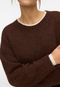 Personne portant un pull texturé marron foncé par-dessus un haut blanc à manches longues, les bras croisés devant un fond uni.