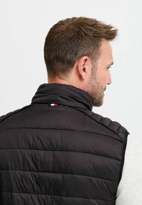 Schwarze Steppweste mit hohem Kragen, horizontalem gestepptem Muster und einem kleinen roten, weißen und blauen Logodetail an der Nackennaht.