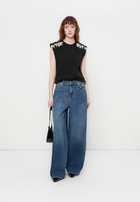 Svart ermeløs topp med utsmykkede skuldre, kombinert med vide blå jeans. Modellen holder en liten svart håndveske med minimalistisk design.