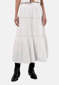 Maxi gonna bianca in cotone con design a strati, dettagli in pizzo e vita elasticizzata. Abbinata a stivali neri in stile cowboy.