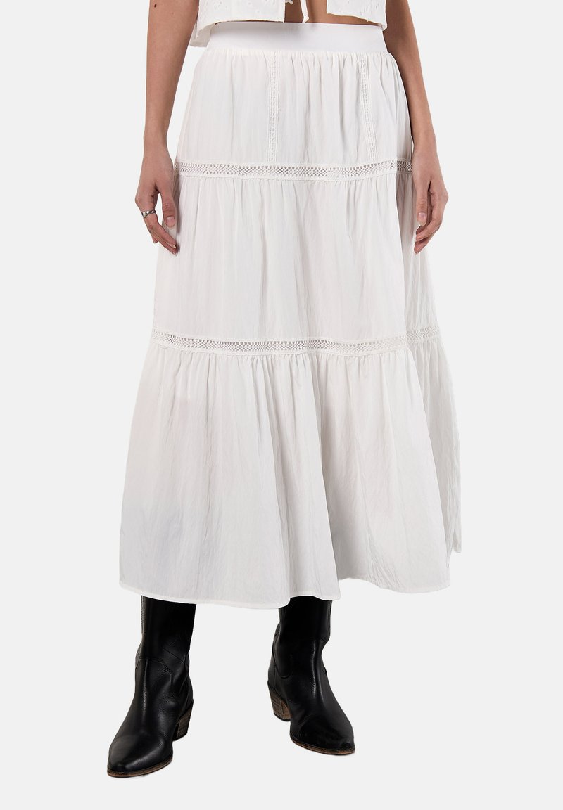 Maxi gonna bianca in cotone con design a strati, dettagli in pizzo e vita elasticizzata. Abbinata a stivali neri in stile cowboy.