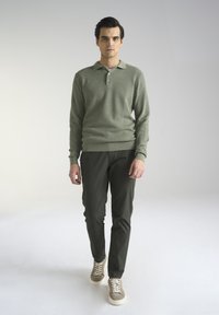 Zielony sweter o teksturowanej fakturze z kołnierzykiem, w połączeniu z ciemnozielonymi spodniami slim-fit oraz beżowymi sneakersami z sznurówkami. Minimalistyczny design, neutralne kolory.