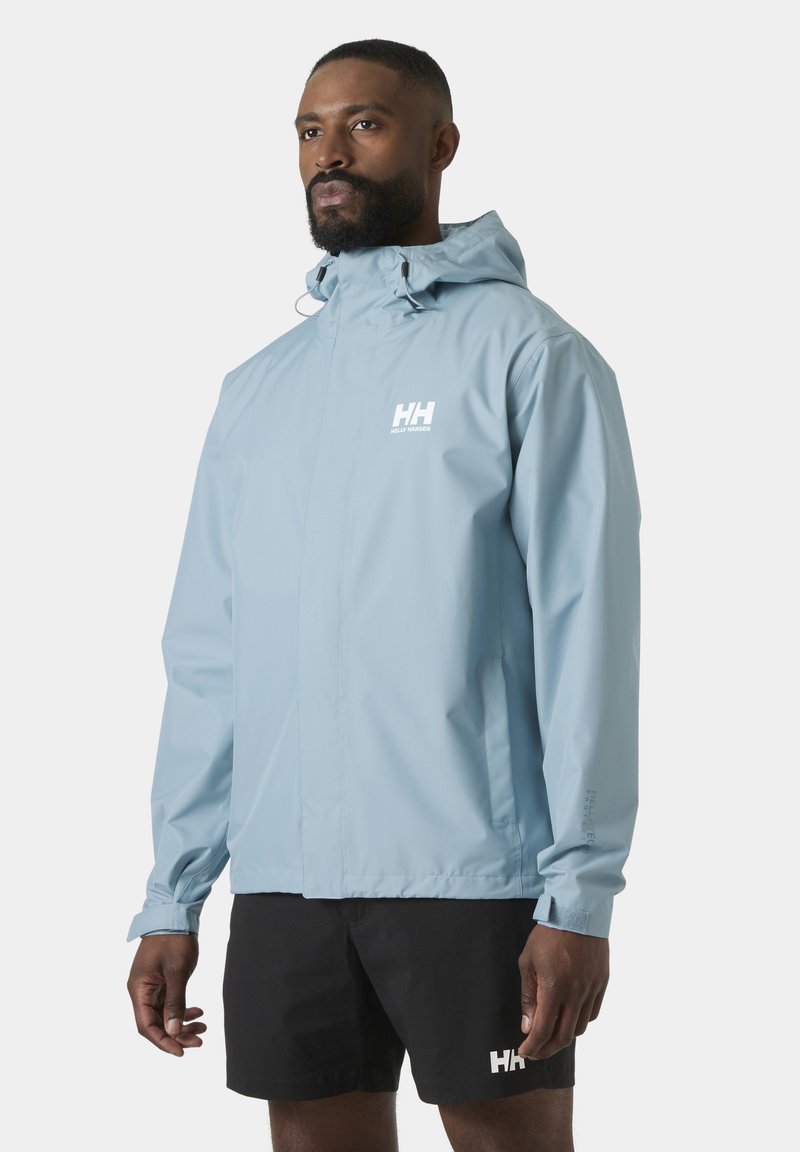 Mann trägt hellblaue wasserdichte Jacke mit Kapuze und schwarze Shorts, steht vor einfarbig hellem Hintergrund.