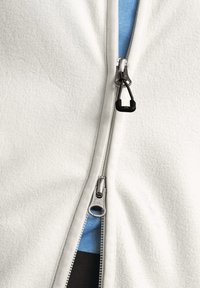 Giacca di pile bianca con zip frontale, caratterizzata da una zip argentata e un tirante nero. Sotto, è visibile una camicia a strati blu e nera.