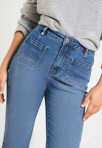 Kõrge vöökohaga sinised denim püksid, mis on keha külge sobiva lõikega, kahte tagakotti ja pruunide õmblustega. Pehme ja sile tekstuur.