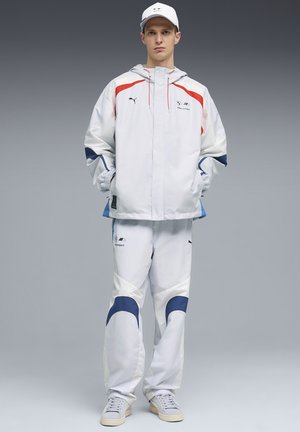 Ung mand iført hvid BMW Motorsport jakke, matchende bukser med blå accenter og hvid kasket, stående mod grå baggrund med hænderne i lommerne.