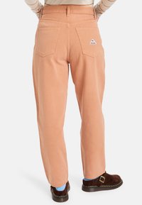 Pantalones de pierna amplia color durazno hechos de denim suave. Presentan dos bolsillos traseros y un pequeño parche con el logo en uno de los bolsillos.