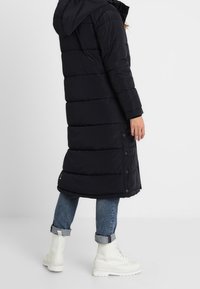 Osoba ubrana w długi czarny pikowany płaszcz z kapturem, niebieskie spodnie jeansowe podwinięte u dołu oraz białe sznurowane botki, stojąca bokiem na białym tle.