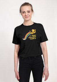 Młoda kobieta w czarnej koszulce z żółtym statkiem wikingów, tęczowym szlakiem i tekstem "Nowe Asgard Tours" z przodu, stojąca na jednolitym tle.