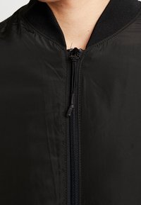 Veste bomber noire avec fermeture éclair à l'avant, col côtelé et tissu lisse et léger. Remarquable pour son design minimaliste et sa texture élégante.