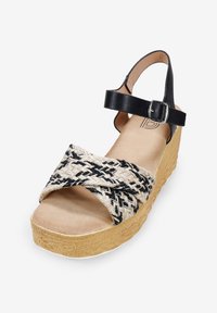 Sandaña de plataforma en negro ybeige con parte superior de tela trenzada, correa de tobillo ajustable con hebilla plateada y suela de cuña de color natural texturizada.