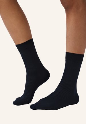 Beine, die schlichte schwarze Sportsocken mit gerippter Struktur tragen, vor einem hellen neutralen Hintergrund stehend.