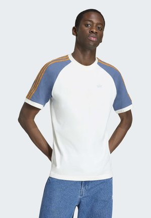 Giovane uomo con le mani dietro la schiena, indossa una maglietta bianca con maniche raglan a righe blu e arancioni e jeans blu, in piedi davanti a uno sfondo chiaro.