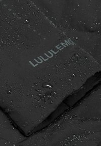 Crna vodootporna jakna u krupnom planu, sa teksturiranom površinom na kojoj su vidljive kapi vode i sivo "LULULEMON" logo na tkanini.
