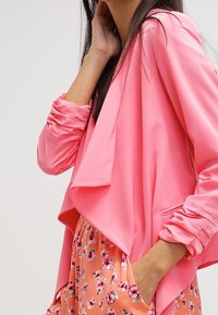 Růžový kabát s dlouhými rukávy, měkkou texturou a otevřeným předním dílem; nošený přes sukně vzorované květinami v oranžové barvě.