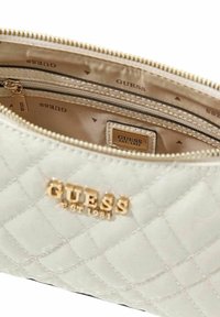 Trousse de maquillage crème matelassée avec ferrures dorées et logo "GUESS EST. 1981". L'intérieur est doublé d'un tissu léger et lisse avec un motif de la marque.