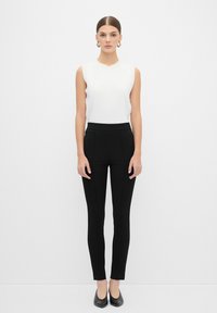 Haut sans manches blanc avec un col rond, associé à un pantalon noir ajusté. Le pantalon présente une taille haute et une texture lisse.