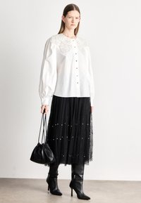 Biela košeľa zapínaná na gombíky s ozdobným čipkovaným golierom, v kombinácii s čiernou čipkovanou sukňou s kruhovými detailmi a čiernymi čižmami po kolená.