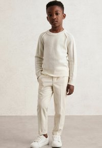 Cremefarbener Strickpullover mit Raglanärmeln, kombiniert mit hellbeigen, taillierten Hosen und weißen Sneakers. Minimalistisches Design, glatte Textur.