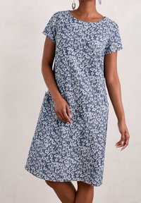 Šaty s krátkým rukávem z modré tkaniny, zdobené bílým květinovým vzorem, s kulatým výstřihem a bočními kapsami. Design je uvolněný a vzdušný.