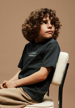 Child with curly hair sits sideways on a padded chair, wearing a dark t-shirt and beige pants, looking to the right against a plain background.