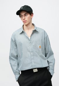 Camisa de rayas, abotonada en azul y blanco, con un bolsillo en el pecho que lleva un logo, combinada con una gorra negra y pantalones negros.