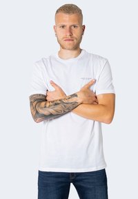 Tricou alb din bumbac cu guler rotund, având un logo mic pe partea stângă a pieptului. Modelul are un braț tatuat și poartă blugi albaștri.