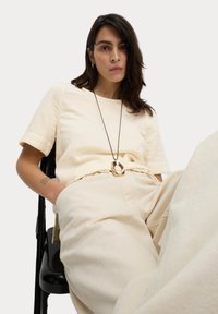 Beige texturerad topp med korta ärmar, tillsammans med höga beige byxor. Ett guldhänge och en grönaktig tatuering på armen.