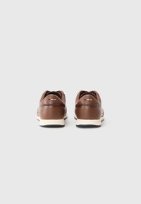 Baskets en cuir marron avec une semelle blanche, présentant une forme arrondie, des coutures minimales et de petites touches de noir et beige sur le talon.