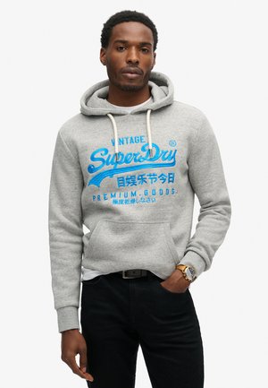 Sudadera gris con un bolsillo tipo canguro, capucha con cordón y texto impreso en azul. Presenta una tela texturizada y puños acanalados.