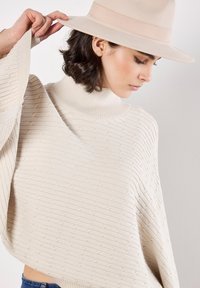 Maglione beige lavorato a maglia con colletto alto e maniche ampie, caratterizzato da un motivo testurizzato. Un cappello a tesa larga di un beige chiaro completa l'outfit.