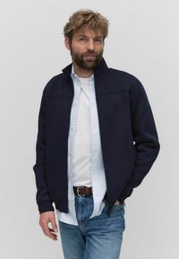 Marinfärgad zip-up jacka med hög krage, tillverkad av mjukt tyg. Bärs över en vit T-shirt och en ljusblå randig skjorta, tillsammans med blå jeans.