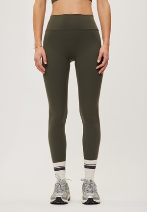 Person iført olivengrønne leggings med høj talje, hvide strømper med striber, grå sneakers, synlig fra midten af maven til fødderne mod en ensfarvet baggrund.