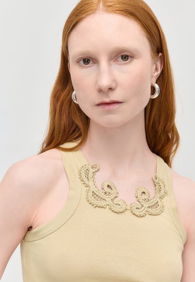 Femme aux longs cheveux roux portant un haut beige sans manches avec des détails en dentelle et des boucles d'oreilles argentées en forme d'anneau, regardant droit devant.