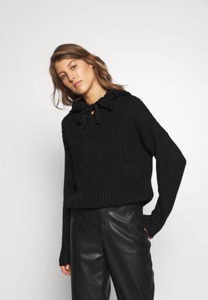 Schwarzer gestrickter Pullover mit Kapuze, ausgestattet mit einem Bindeband am Hals, weiten Ärmeln und gerippter Textur. Kombiniert mit schwarzen Lederhosen.