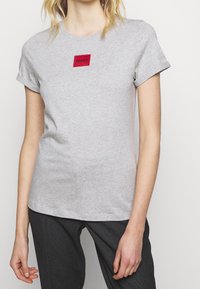 Grå bomulds T-shirt med et rødt rektangulært logo-patch med teksten "HUGO". Korte ærmer og rund halsudskæring, afslappet pasform.