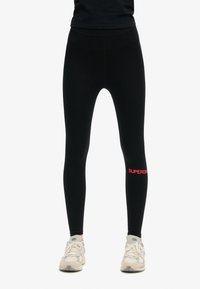 Leggings neri con una texture liscia, caratterizzati da un logo "SUPERDRY" rosso sulla gamba inferiore. Abbinati a scarpe da ginnastica di colore chiaro.