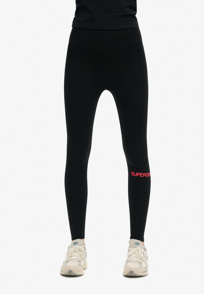 Leggings neri con una texture liscia, caratterizzati da un logo "SUPERDRY" rosso sulla gamba inferiore. Abbinati a scarpe da ginnastica di colore chiaro.
