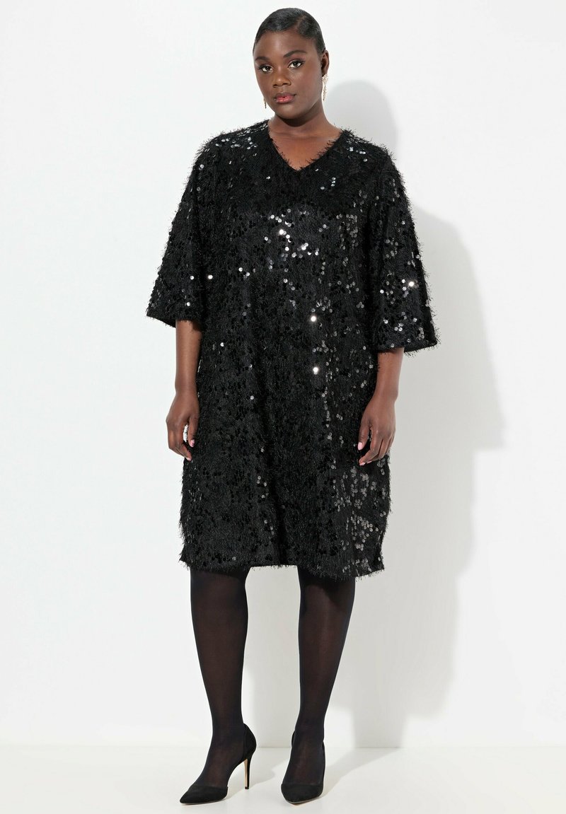 Schwarzes oversized Kleid mit kurzen Ärmeln, aus einem glänzenden Stoff mit strukturierten Oberflächen. V-Ausschnitt-Design. Kombiniert mit schwarzen Strumpfhosen und Absatzschuhen.