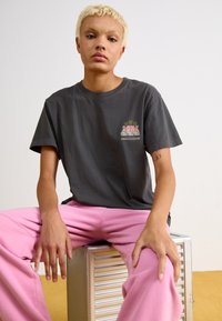 Grå bomull T-skjorte med grafisk design som viser palmer, kombinert med lys rosa bukser. Modellen sitter på en sølvmetallisk overflate.