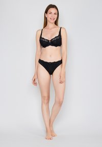 Zwarte kanten bh en bijpassende string met verfijnde kanten afwerking, zachte stof, beugelondersteuning en een klassieke snit. Model poseert rechtop.
