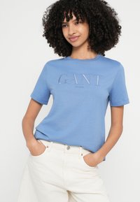 Világoskék pamut póló kerek nyakkal és rövid ujjakkal. Elöl hímzett "GANT" logó díszíti, krémszínű nadrággal párosítva.