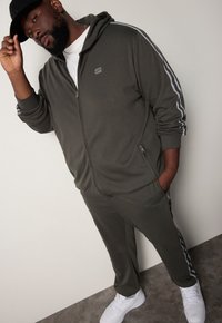Olivengrøn zip-up hoodie med grå striber, matchende olivenfarvede bukser og hvide sneakers. Glat stof, sidelommer, afslappet pasform.