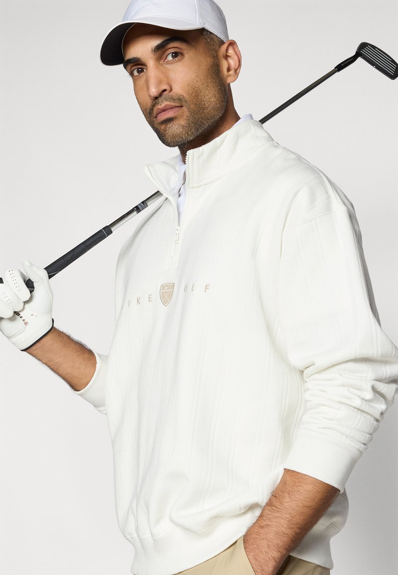 Weißer Golfpullover mit Reißverschluss und strukturierten Streifen, mit einem auf der Brust platzierten Logo und gerippten Bündchen, getragen mit einer weißen Mütze und einem Golfschläger.