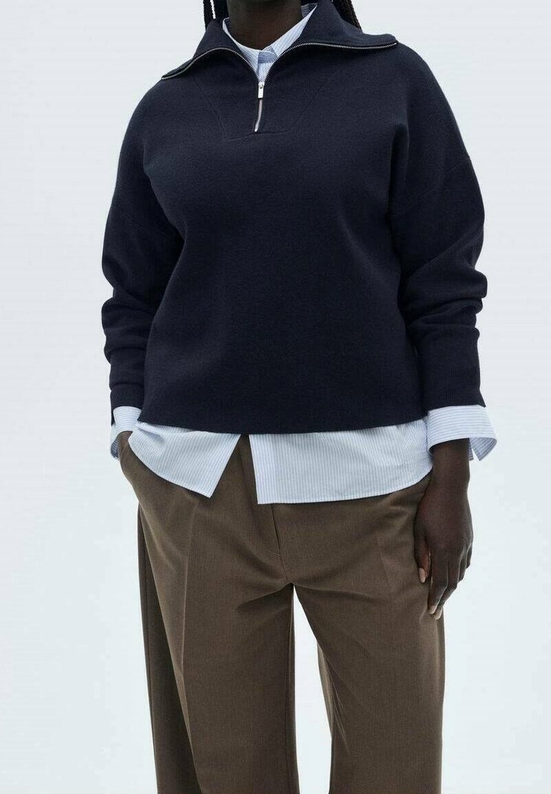 Personne portant un pull à fermeture éclair quart-de-tour bleu marine superposé à une chemise blanche rayée, et un pantalon marron taille haute, les mains dans les poches.