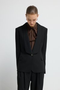 Zwart blazer met krijtstreep, brede revers en sluiting met een enkele knoop, gecombineerd met een bruine blouse met een strik bij de hals. Model staat met het hoofd naar beneden.