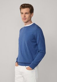 Niebieski sweter robiony na drutach z okrągłym dekoltem, długimi rękawami i ściągaczami, sparowany z białymi spodniami. Gładka tekstura, bez wzorów.